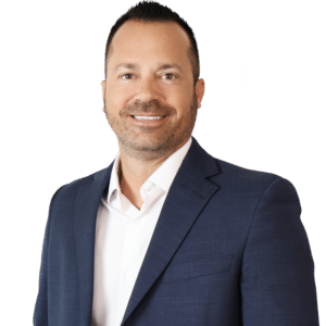 Man wearing a navy blue suit jacket and white shirt, smiling, standing against a plain white background.