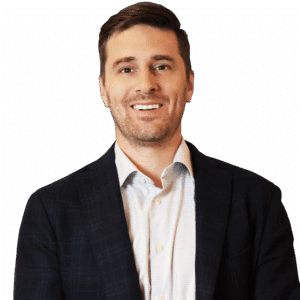 A man wearing a dark blazer over a light-colored shirt is standing and smiling at the camera against a white background.
