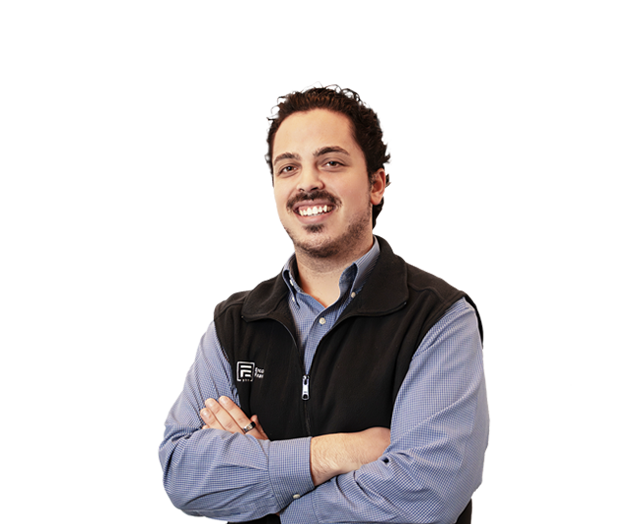 Man with dark hair and a beard, wearing a blue shirt and black vest, standing with arms crossed and smiling against a plain white background.