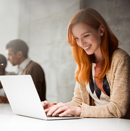 A smiling woman with red hair using a laptop in an office setting, with two colleagues in the background.