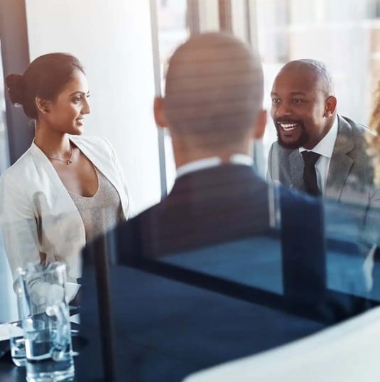 Group of professionals in a meeting room, engaged in conversation, with two visible through glass reflections.