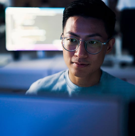 Person wearing glasses looking at a computer screen in a dimly lit room. Background shows blurred computer monitors with code.