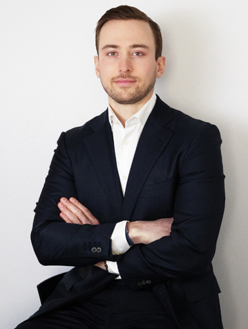 A man in a dark suit and white dress shirt sits with arms crossed, facing forward against a plain white background.