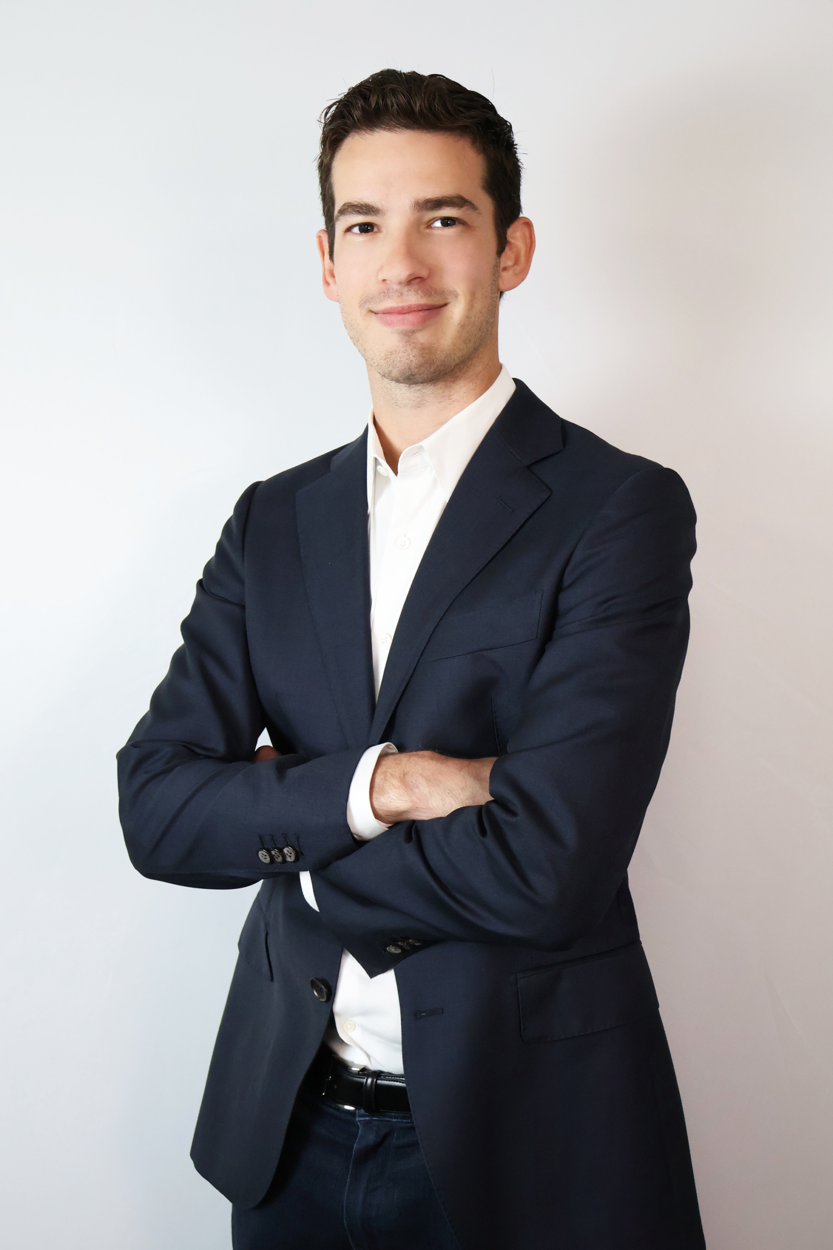 A man in a dark suit and white shirt stands with arms crossed, facing the camera against a plain light background.
