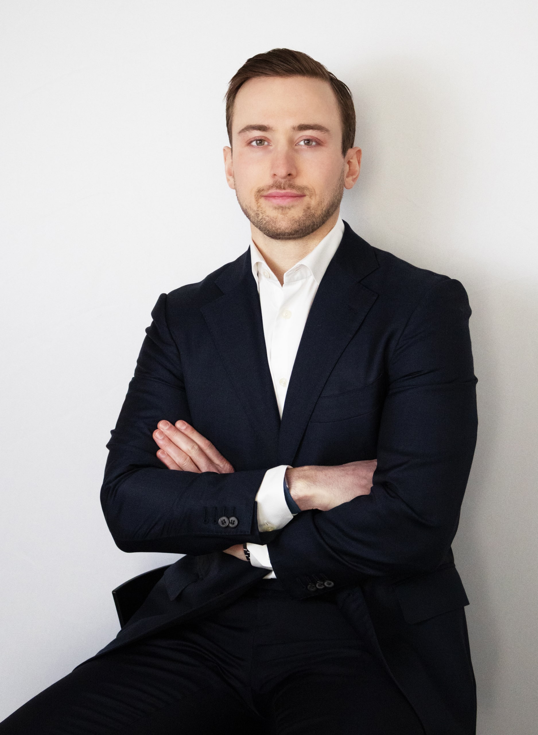 A man in a dark suit and white shirt sits against a plain background with arms crossed, looking at the camera.