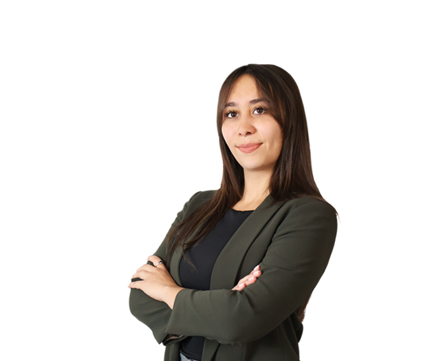 A woman with long brown hair wearing a dark blazer stands with arms crossed, looking ahead with a neutral expression against a white background.