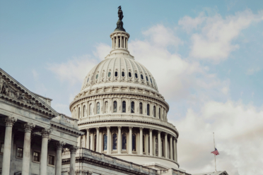 The dome of the United States Capitol building is shown against a partly cloudy sky, with part of the American flag visible in the foreground.