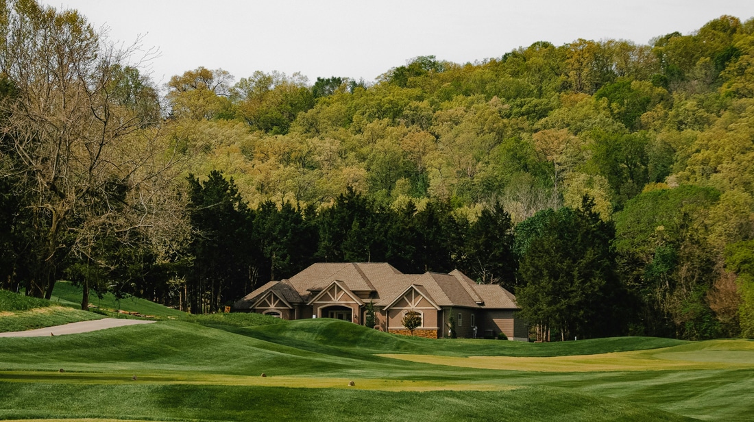 A large house sits among trees and hills, with neatly mowed grass and a paved path in the foreground.