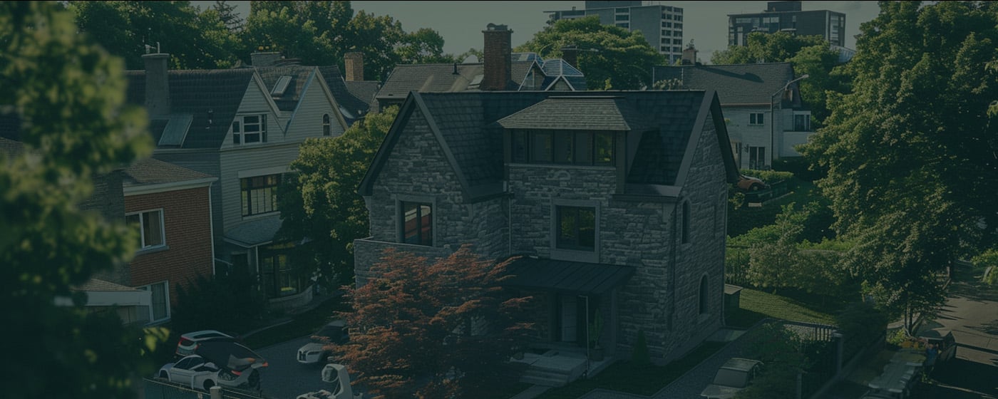 A stone house with a dark roof sits among other houses on a tree-lined residential street, with several cars parked nearby.