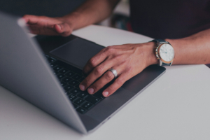 A person wearing a watch and ring types on a laptop at a white table.