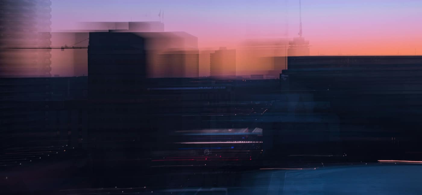 Blurred city skyline at sunset with darkened buildings and a gradient sky transitioning from orange to purple tones.