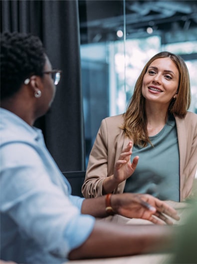 Two people sit at a table having a conversation in a modern office setting. One woman gestures while speaking, and the other listens attentively.