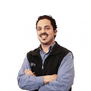 Man with dark hair and facial hair, wearing a blue shirt and black vest, standing with arms crossed and smiling against a plain white background.
