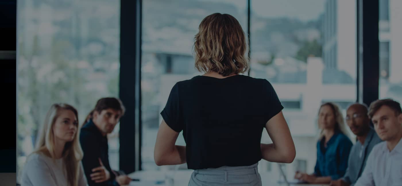 A woman stands and speaks to a group of seated colleagues in a modern office conference room with large windows.