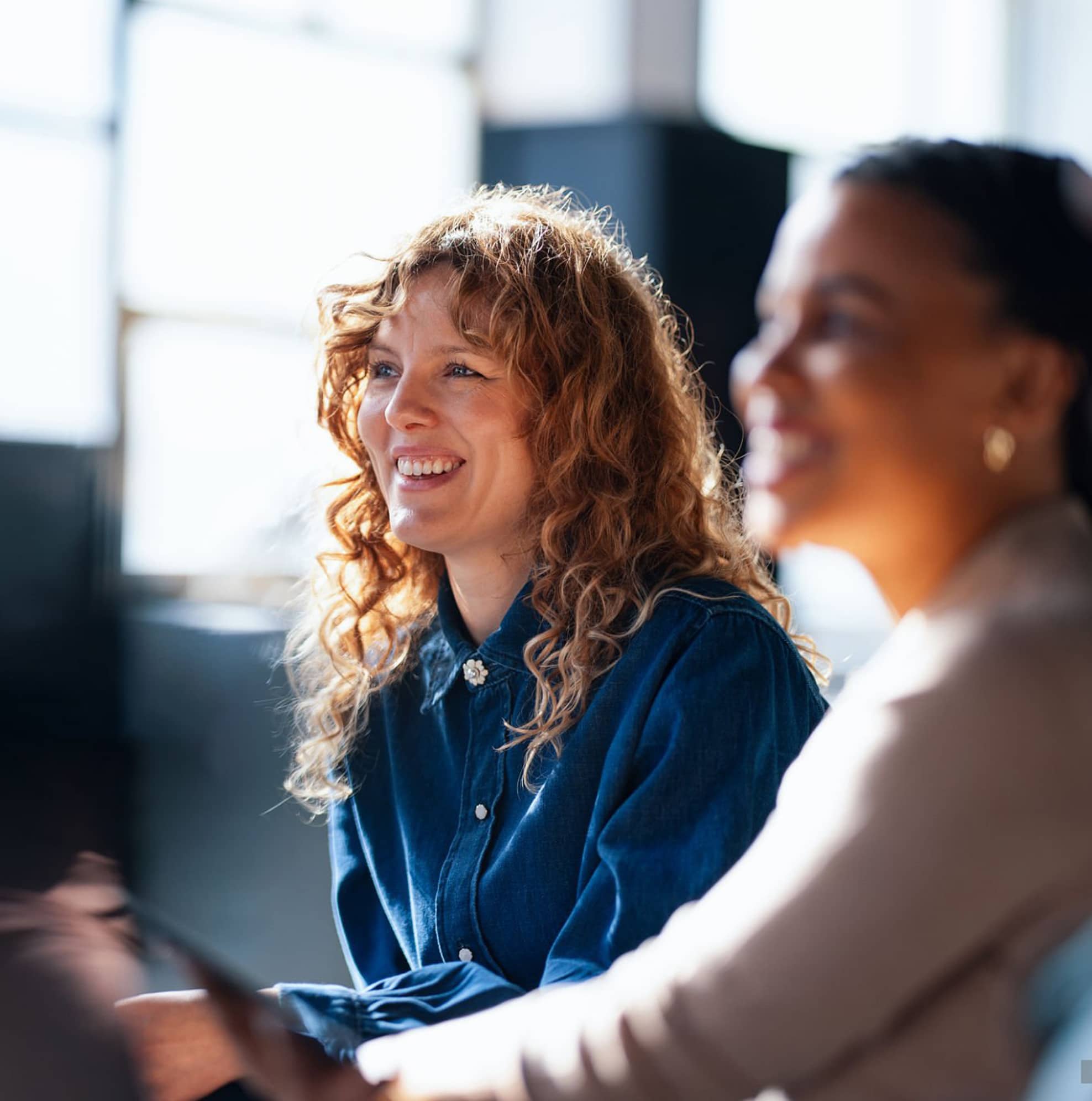 Two women sit side by side indoors, smiling and looking in the same direction, with sunlight streaming in from a nearby window.