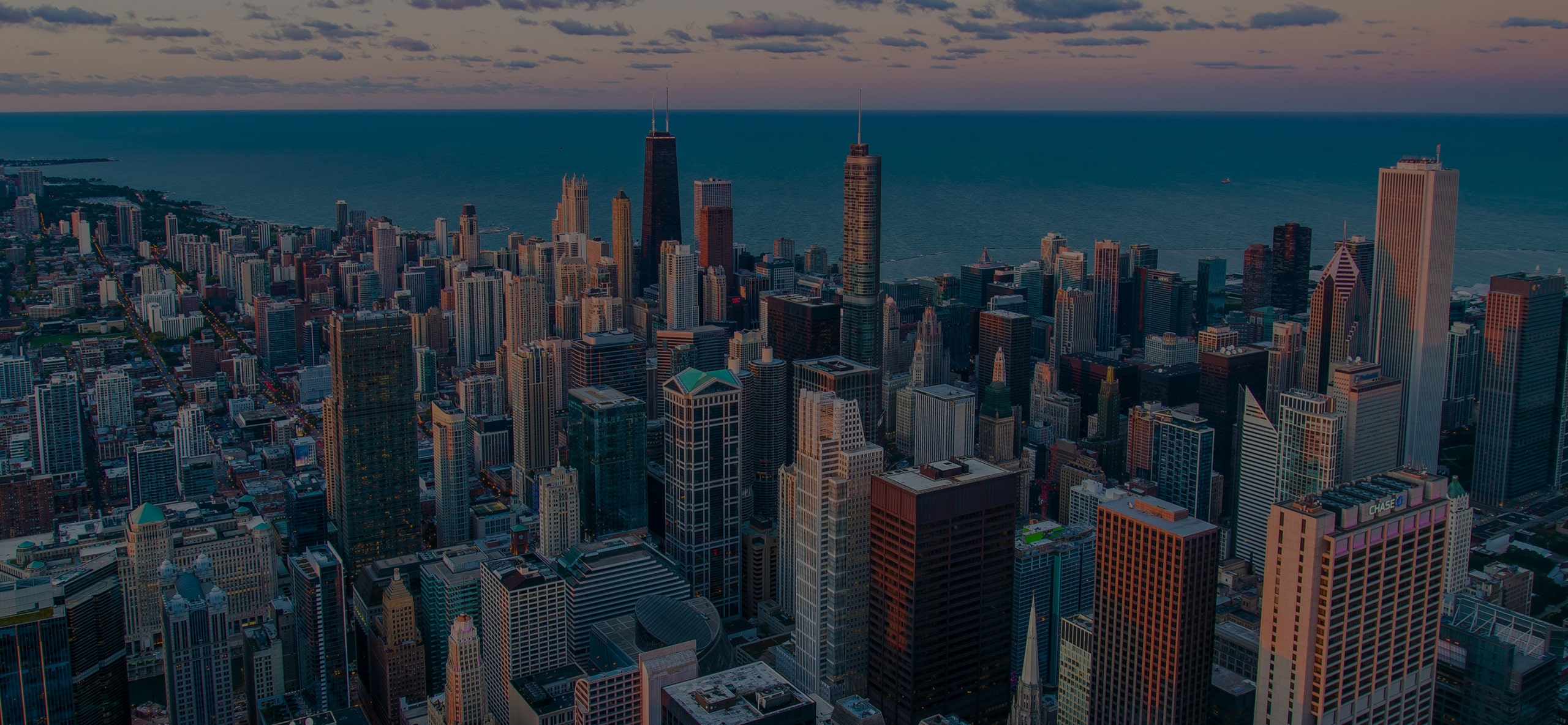 Aerial view of a city skyline with tall skyscrapers near a large body of water under a partly cloudy sky at sunset.