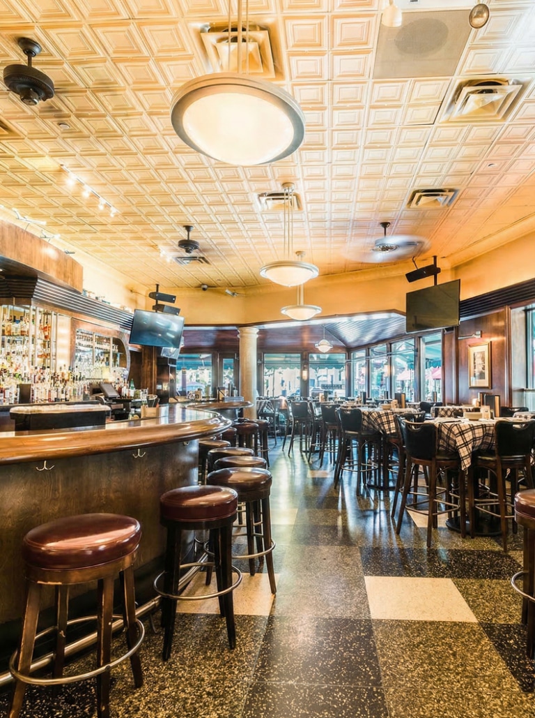 Spacious restaurant interior with a bar, high stools, tables set for dining, checkered floor, large windows, and overhead lighting.