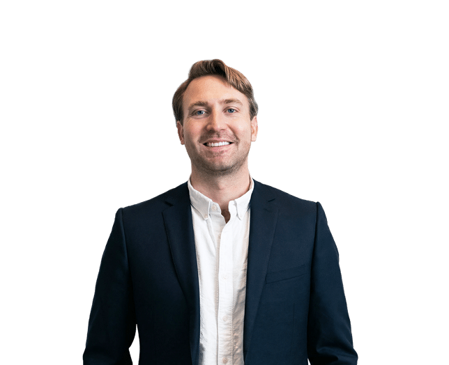 Man wearing a dark suit jacket and white shirt stands against a plain white background, smiling at the camera.