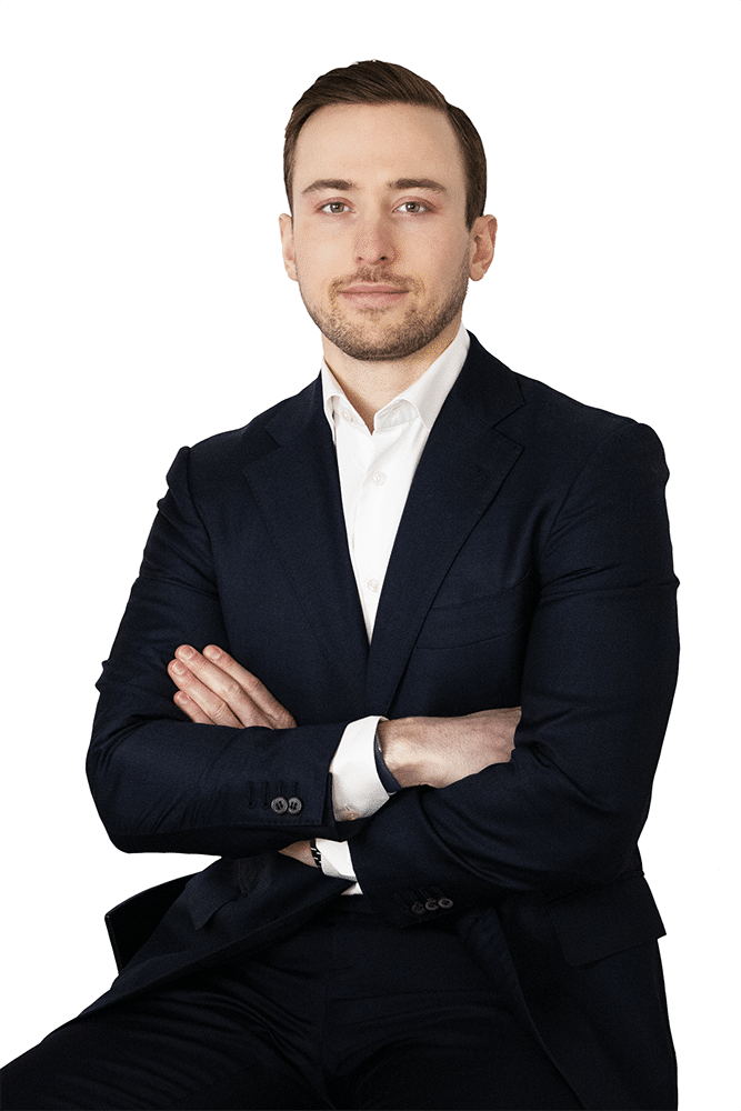 Man in a dark suit and white shirt sitting with arms crossed, facing forward against a plain white background.