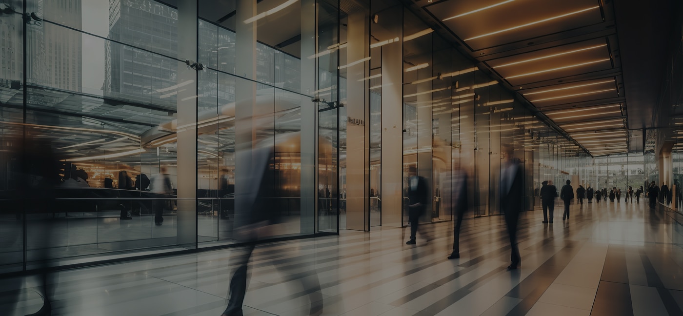 People walk through a modern glass building lobby with blurred motion, featuring bright lights, tall windows, and reflections on the polished floor.