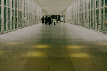 A spacious, brightly lit corridor with glass walls and a tiled floor, showing several people walking away into the distance.