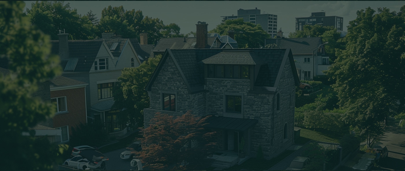 A stone house with a dark roof is surrounded by trees and neighboring houses in a residential area, with apartment buildings visible in the background.