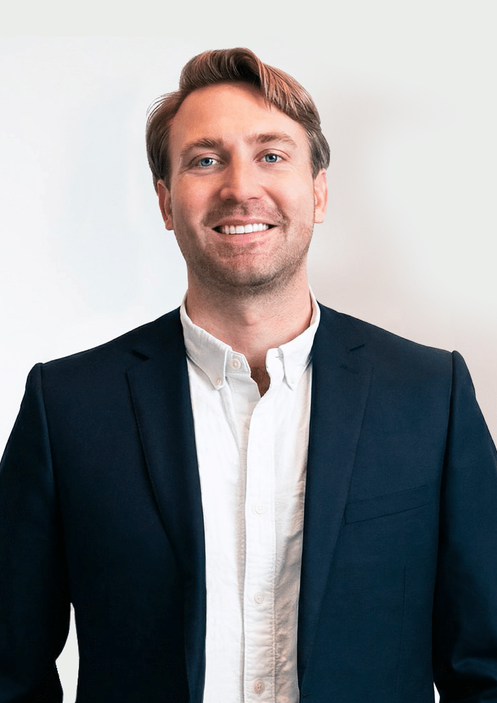 A man in a dark suit jacket and white shirt stands against a plain light background, smiling at the camera.