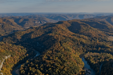 Aerial view of rolling hills covered in autumn foliage with winding rivers and scattered clearings under a partly cloudy sky.