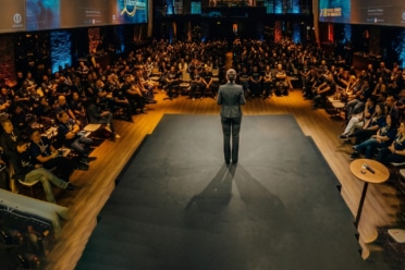 A speaker stands on a stage addressing a large audience seated in a dimly lit auditorium with screens displaying presentation slides overhead.