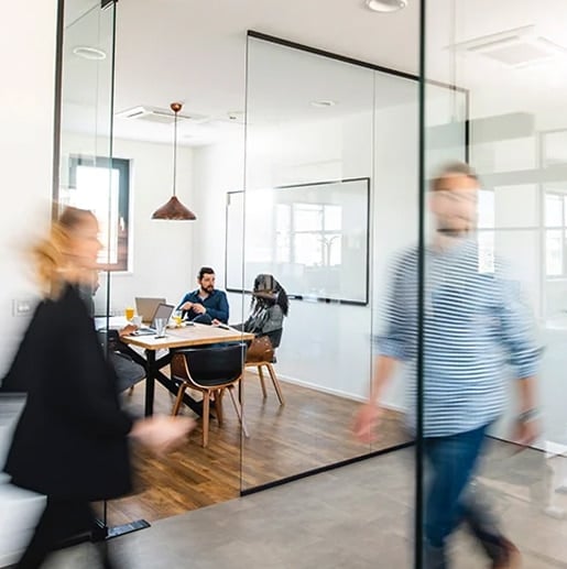 Two people walk past a glass-walled office where three people are seated at a table with laptops and papers, engaged in a meeting.
