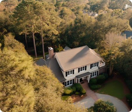 Aerial view of a two-story house with a gray roof, surrounded by trees and greenery, with a driveway and brick pathway leading to the entrance.