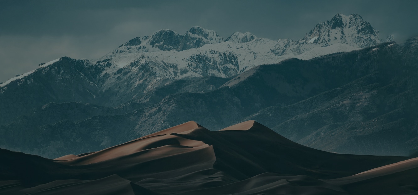 Dark sand dunes in the foreground with snow-capped mountains in the background under a cloudy sky.