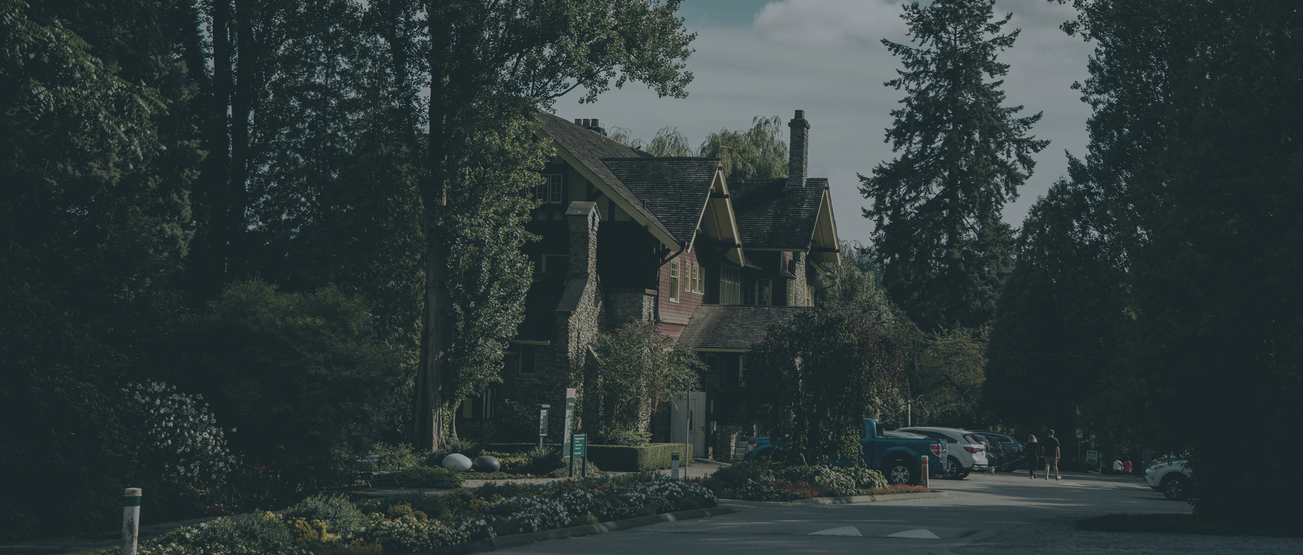 A large house surrounded by trees and landscaped gardens, with several parked cars and a partly cloudy sky overhead.