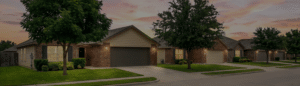 Row of single-story suburban homes with brick and siding exteriors, green lawns, trees, and driveways at sunset.