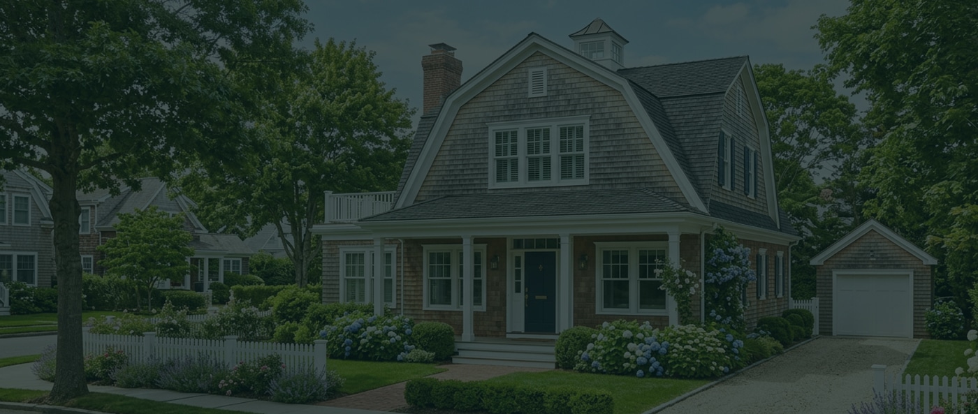 A two-story suburban house with a front porch, manicured lawn, garden, white picket fence, and detached garage, surrounded by trees.