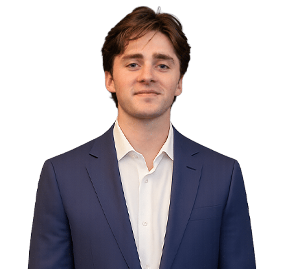 A young man with brown hair wearing a blue suit jacket and white shirt stands in front of a plain white background, looking at the camera.