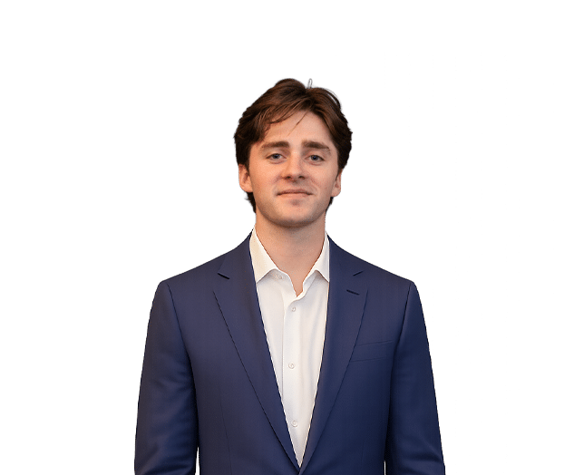 A young man with brown hair wearing a blue suit and white shirt stands against a plain white background.
