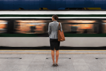 A woman stands on a train platform facing a moving train, with the station floor and blue-tiled wall visible in the background.
