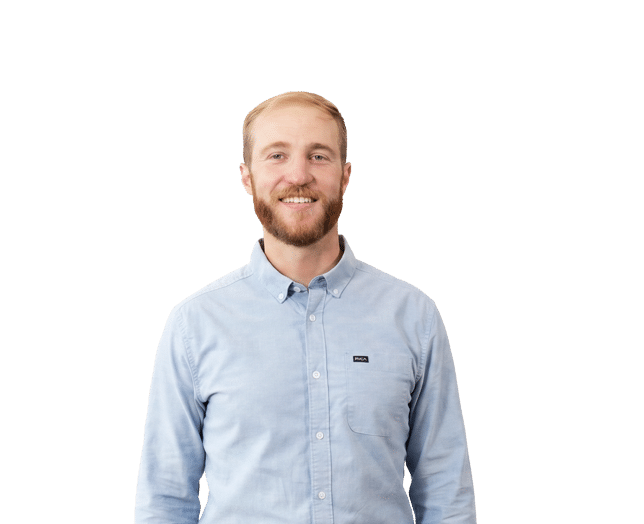 A man with a beard and light brown hair, wearing a light blue button-up shirt, stands against a plain white background, smiling at the camera.