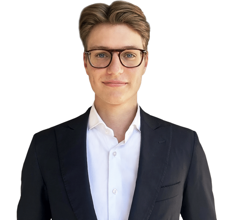 A man wearing glasses, a white shirt, and a black blazer stands against a plain white background, facing the camera.