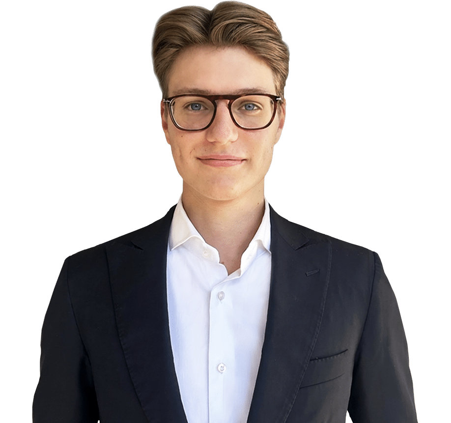 A man wearing glasses, a white shirt, and a black blazer stands against a plain white background, facing the camera.
