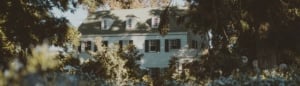 A three-story white house with green shutters is partially obscured by trees and shrubs on a sunny day.