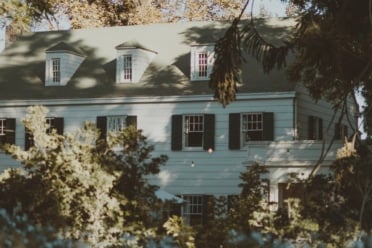 A three-story white house with green shutters is partially obscured by trees and shrubs on a sunny day.