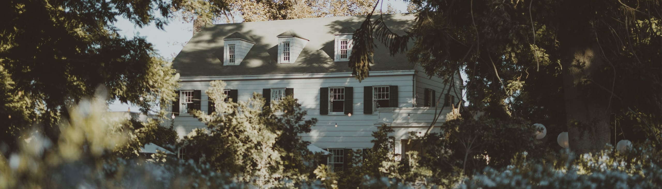 A three-story white house with green shutters is partially obscured by trees and shrubs on a sunny day.