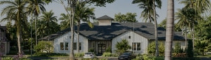 Single-story white house with a dark roof, surrounded by palm trees and greenery; two cars are parked in the driveway on a sunny day, capturing the charm and allure of Florida real estate.