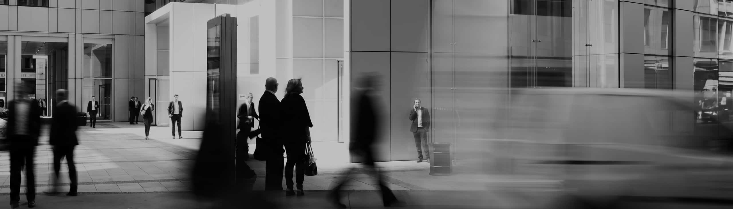 People walking in front of modern office buildings, some in focus and others blurred in motion. Urban street scene with business attire and a van passing by, capturing the energy of a city that thrives on financing through every stage of an investment.