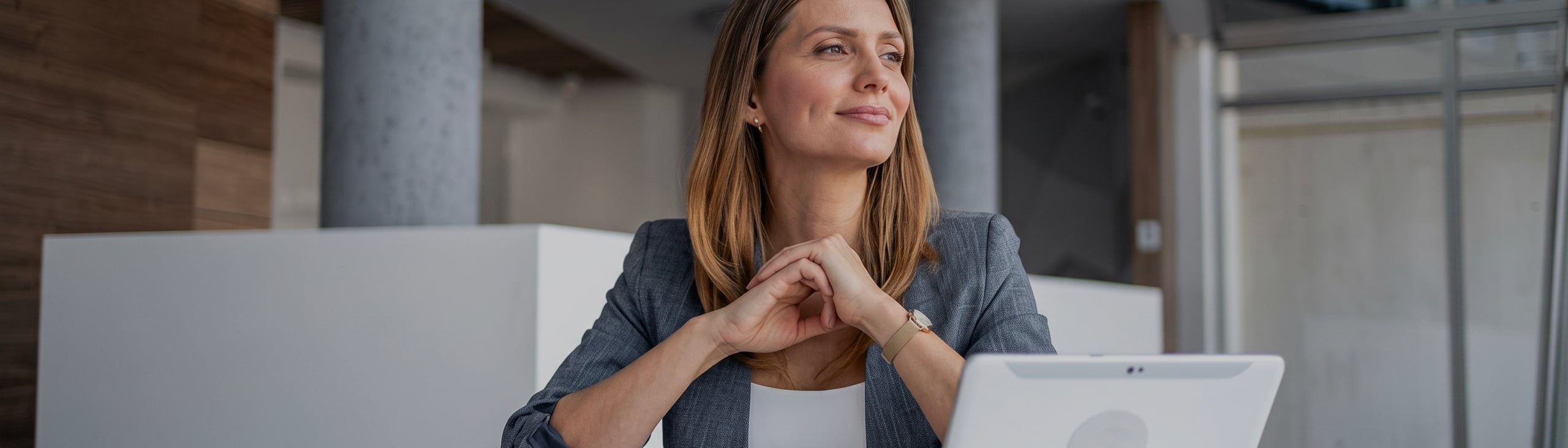 Woman in a gray blazer sits at a desk with a tablet, looking thoughtfully to the side in a modern office setting—perhaps considering how to prep your property for a smooth appraisal.