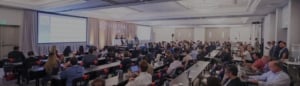 Conference attendees sit at tables in a large, modern room, facing a stage where several speakers present information on a screen about Financing Through Every Stage of an Investment.