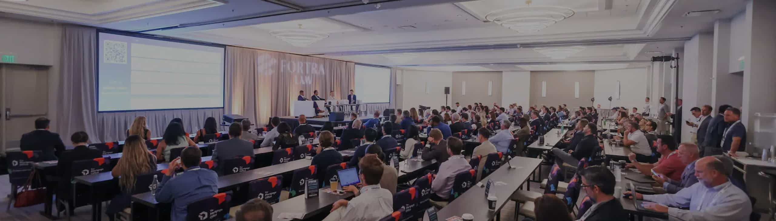Conference attendees sit at tables in a large, modern room, facing a stage where several speakers present information on a screen about Financing Through Every Stage of an Investment.