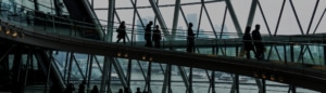 People walking on an elevated walkway inside a modern glass building with a cityscape and river visible through the windows in the background, echoing the atmosphere of insightful events like What I Heard at FORTRA: Why Your Lender Matters in Today’s Market.